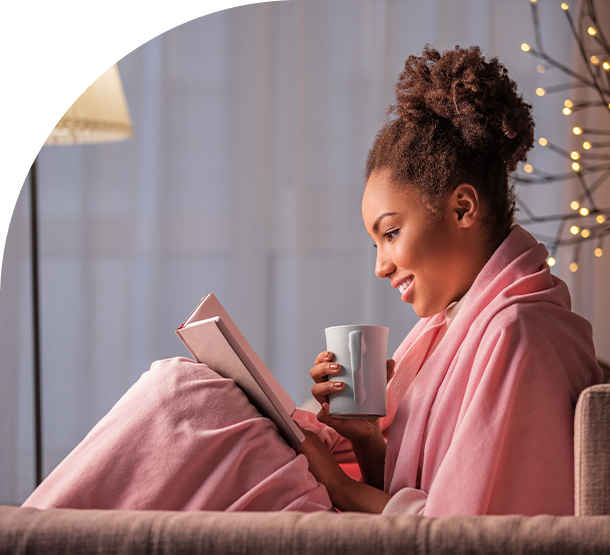 A woman in pink robe reading a book and drinking coffee.